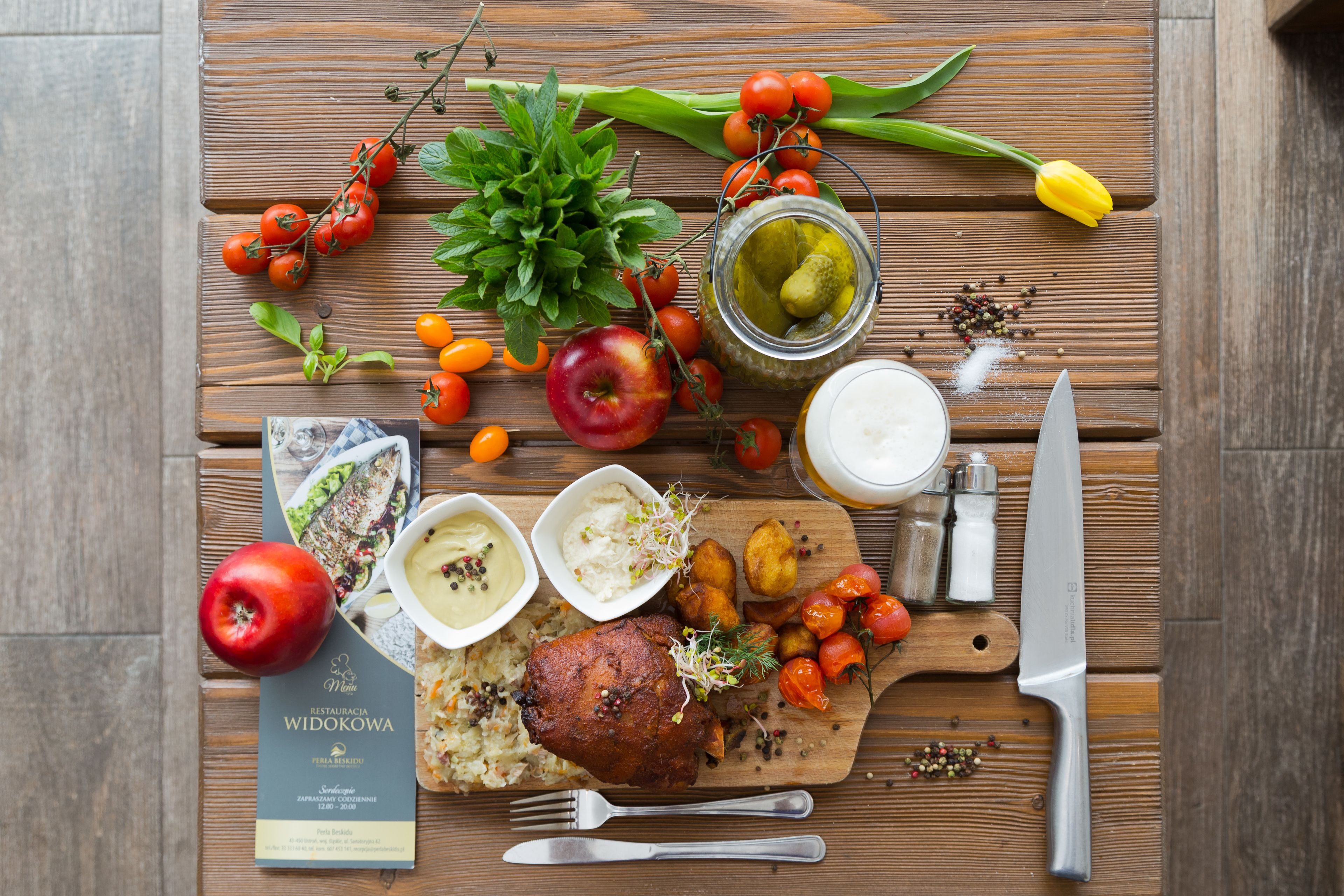 A wooden table with a variety of food items including a red apple, cherry tomatoes, a yellow tulip, and a jar of pickles.