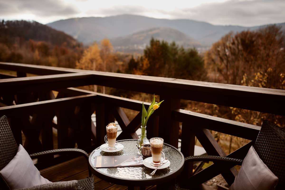Una mesa con dos bebidas y un florero con una flor en un balcón con vistas a un paisaje montañoso.