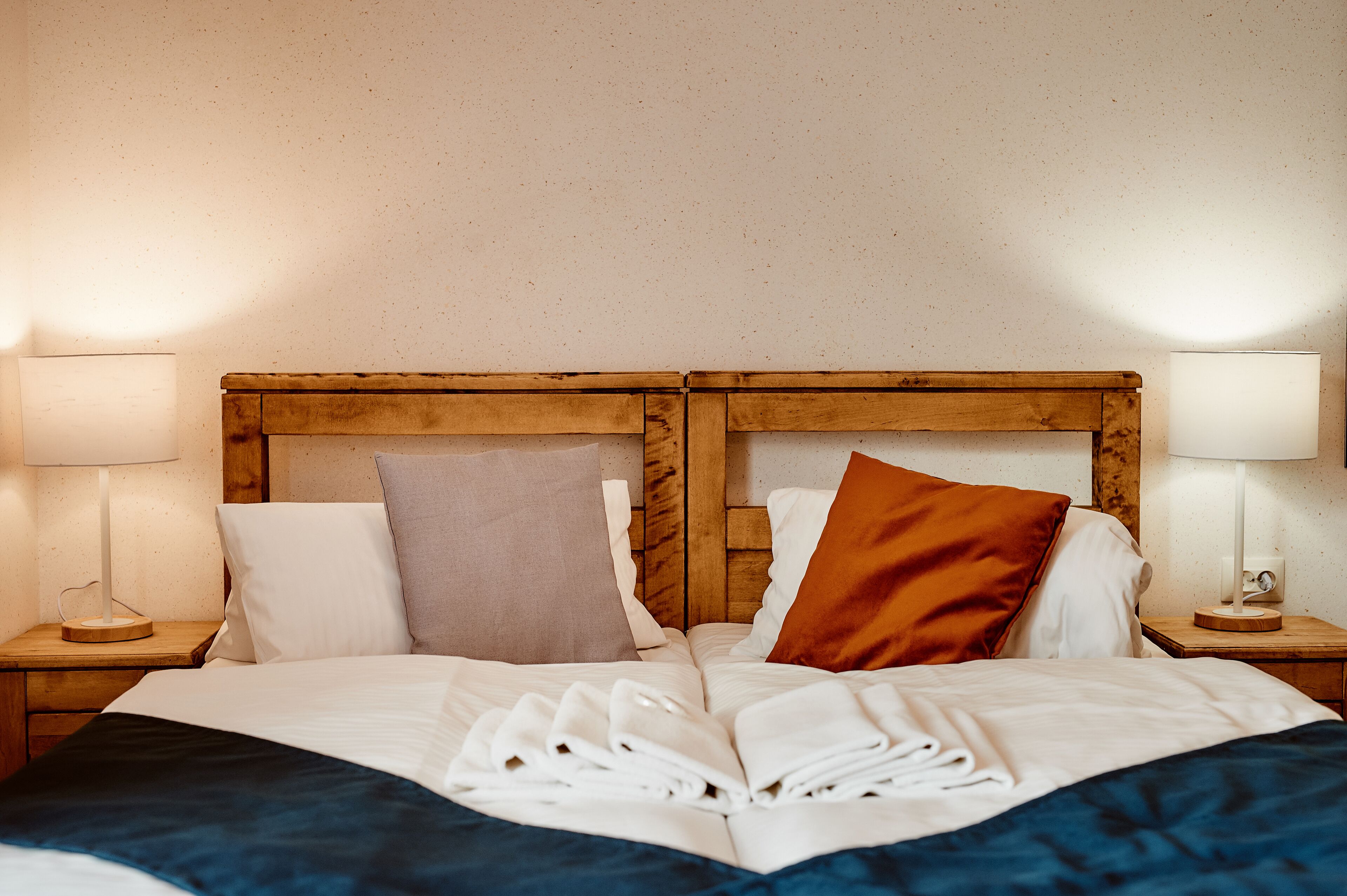 Una cama bien tendida con ropa de cama blanca y azul, dos mesitas de noche de madera con lámparas y un cabecero de madera.