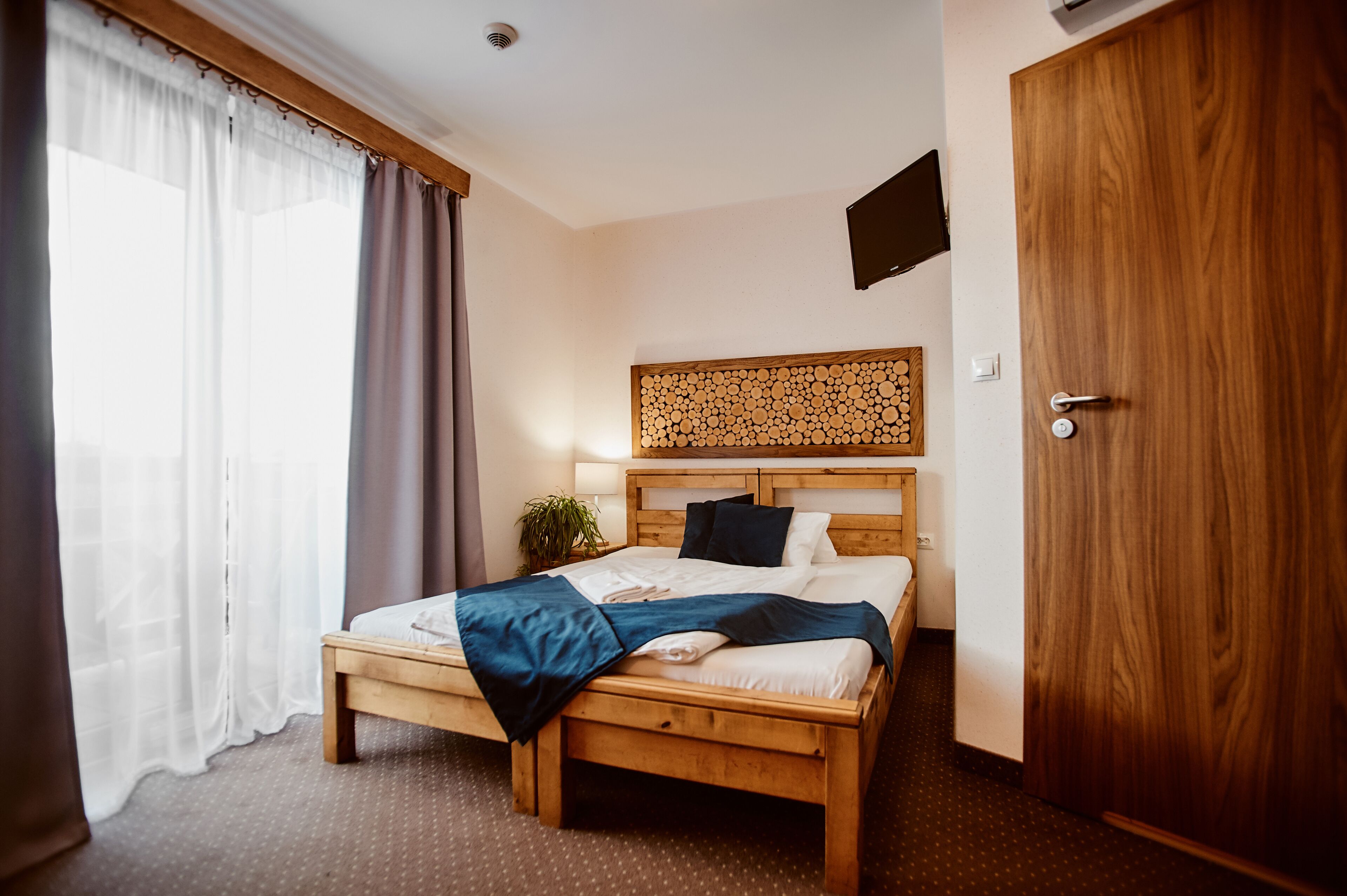 Una cama bien tendida con cabecera de madera, una planta en la mesita de noche y un televisor de pared.