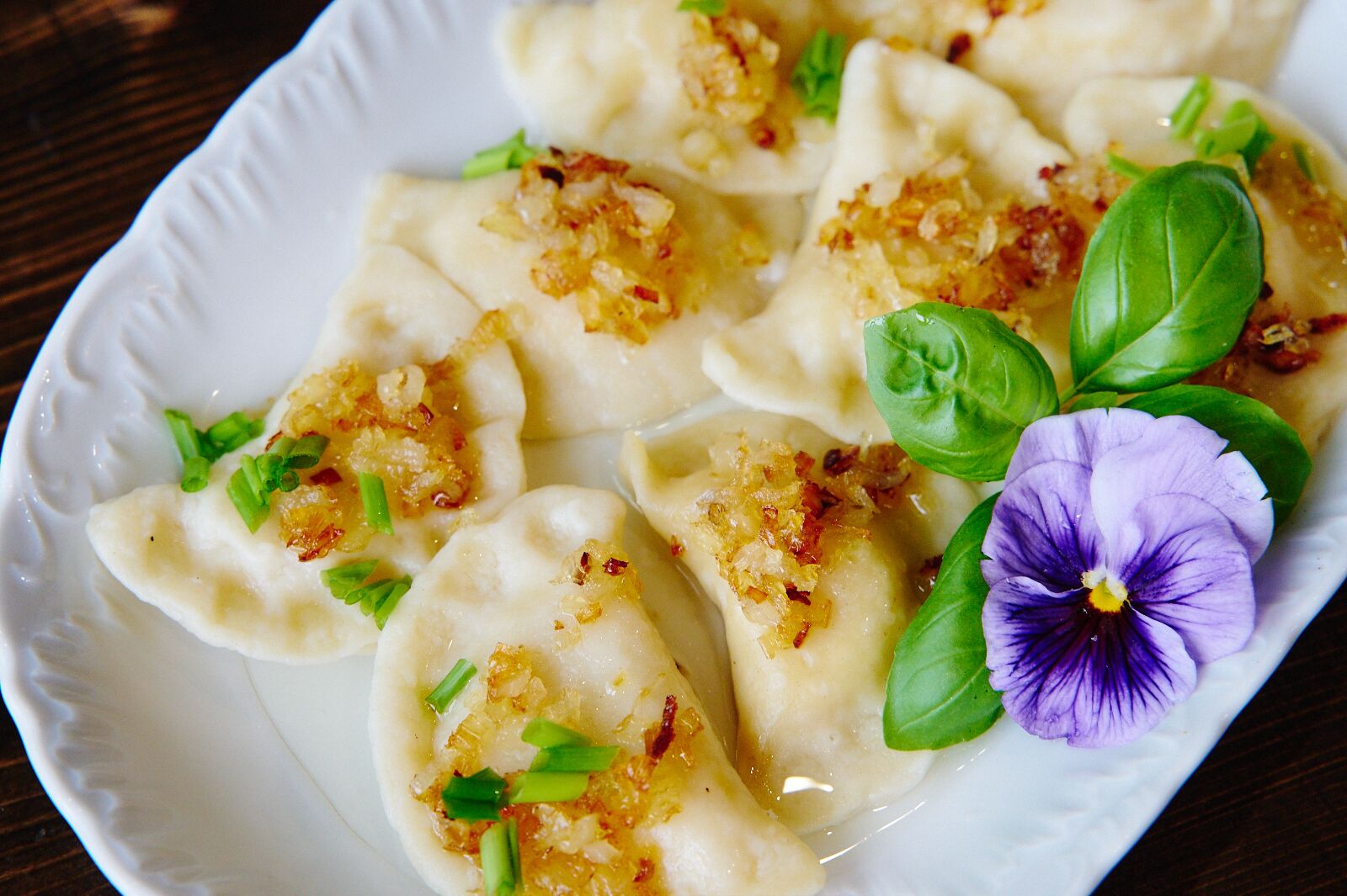 A plate of dumplings garnished with green onions and fried onions, accompanied by a basil leaf and a purple flower.