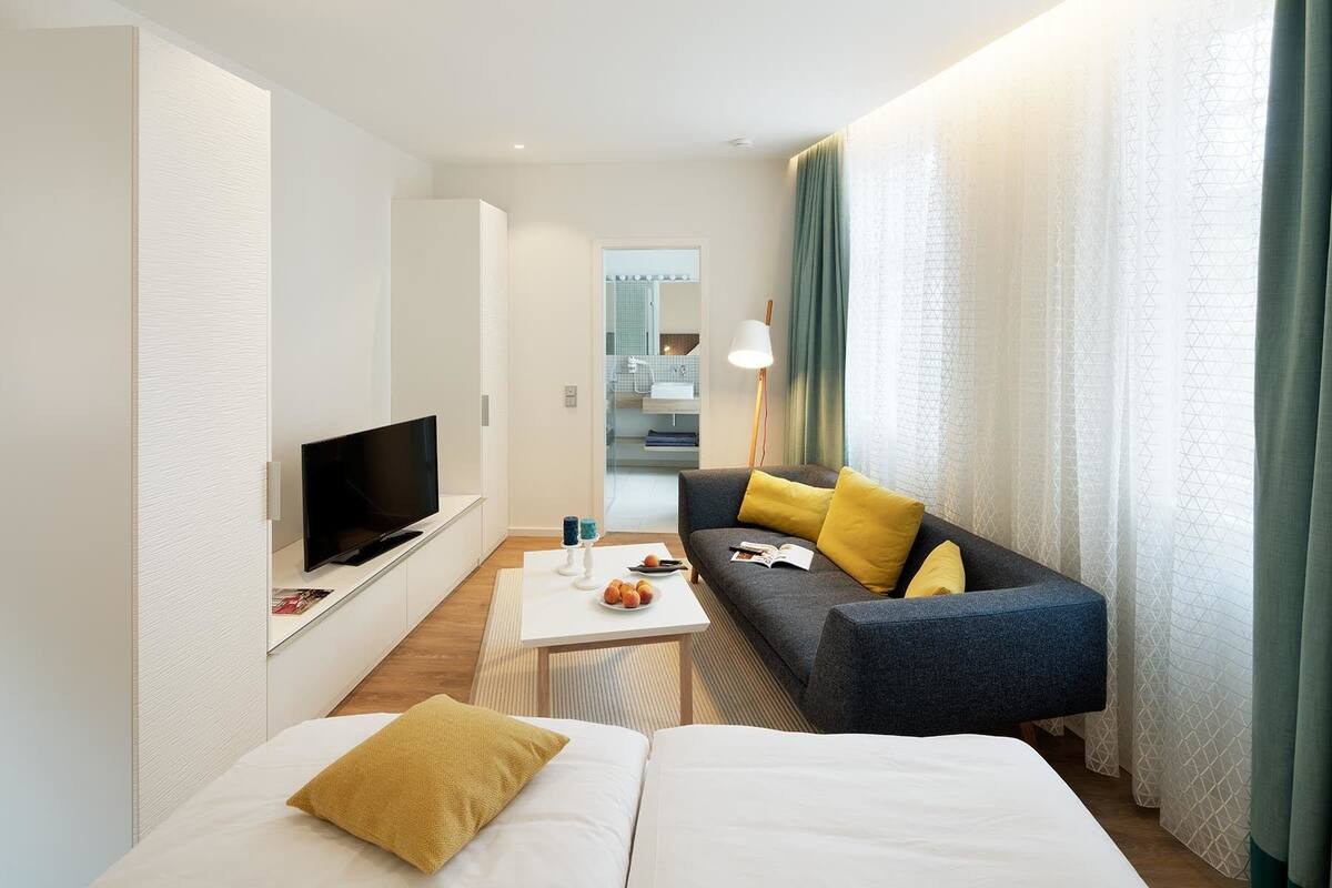 A modern hotel room with a flat-screen TV, a sofa with yellow cushions, a white bed, and a small table with a bowl of fruit.