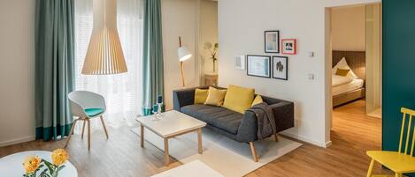 Una sala moderna con un sofá, una mesa de centro y sillas. Se observa una mesa de comedor con una revista y vasos. Una cama es visible al fondo.