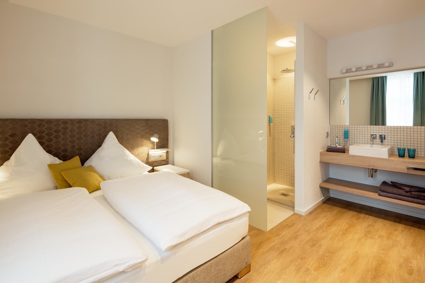 Habitación de hotel con una cama grande, una mesita de noche, un baño con lavamanos y espejo, y piso de madera.