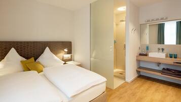 Habitación de hotel con una cama grande, una mesita de noche, un baño con lavamanos y espejo, y piso de madera.
