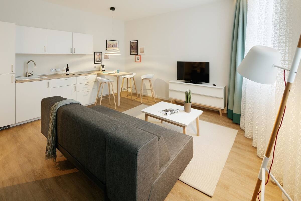 A modern living room with a grey sofa, a dining area with stools, a kitchen with white cabinets, a TV, and a large window with curtains.
