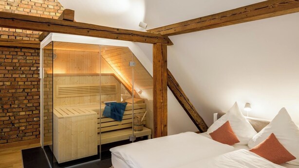 A modern bedroom with a wooden sloped ceiling, a glass-enclosed sauna, and a bed with white and orange pillows. - Goldkind Feine Wohn & Schlafstuben (Heppenheim)