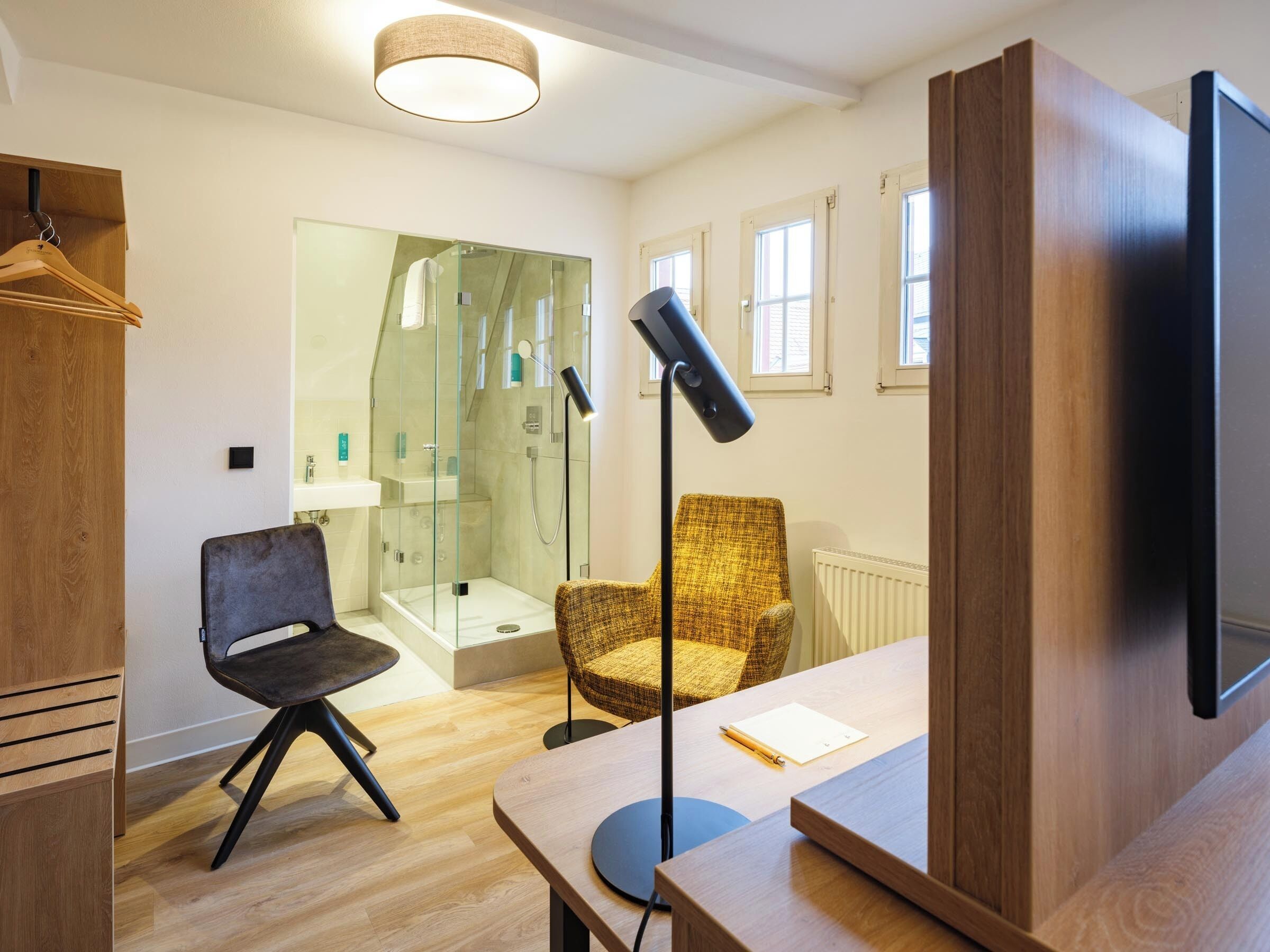 A modern bathroom with a glass shower enclosure, a wooden vanity, a black chair, a yellow armchair, and a desk with a lamp.