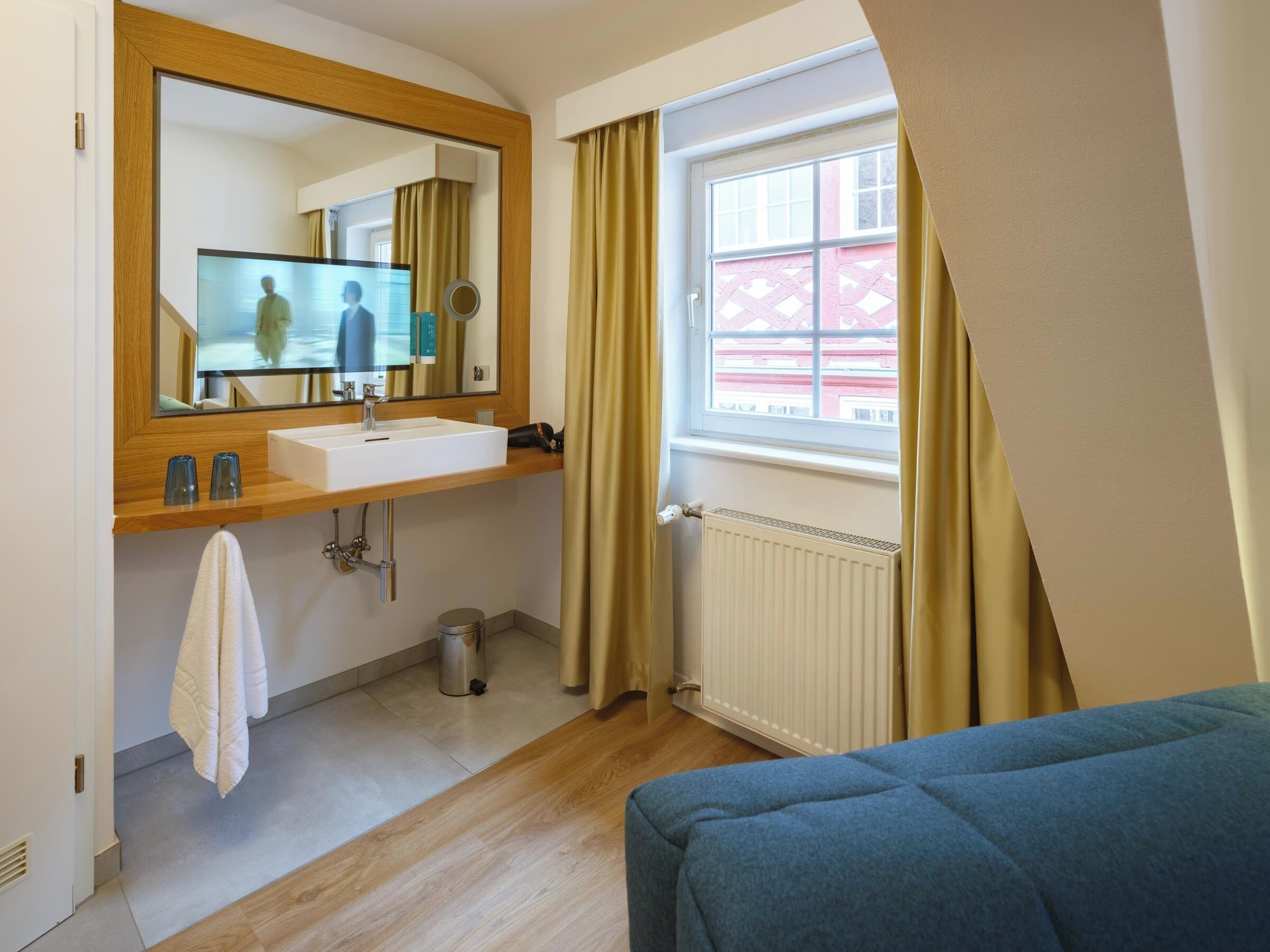 Een moderne badkamer met een houten wastafelmeubel, een grote spiegel, een raam met gordijnen, een radiator en een blauwe bank in de hoek.