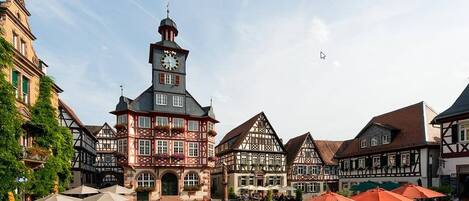 Una plaza tradicional de pueblo europeo con edificios de entramado de madera, una destacada torre del reloj y zonas de estar al aire libre.