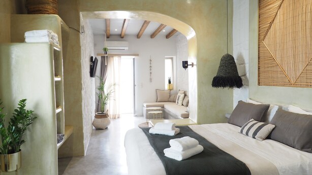 A modern bedroom with a large bed, a built-in wardrobe, a seating area, and a view through an archway. - Aegis Royale Villa (Paros)