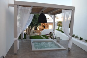 Un área de spa con hidromasaje, paredes blancas y una pérgola con cortinas blancas.