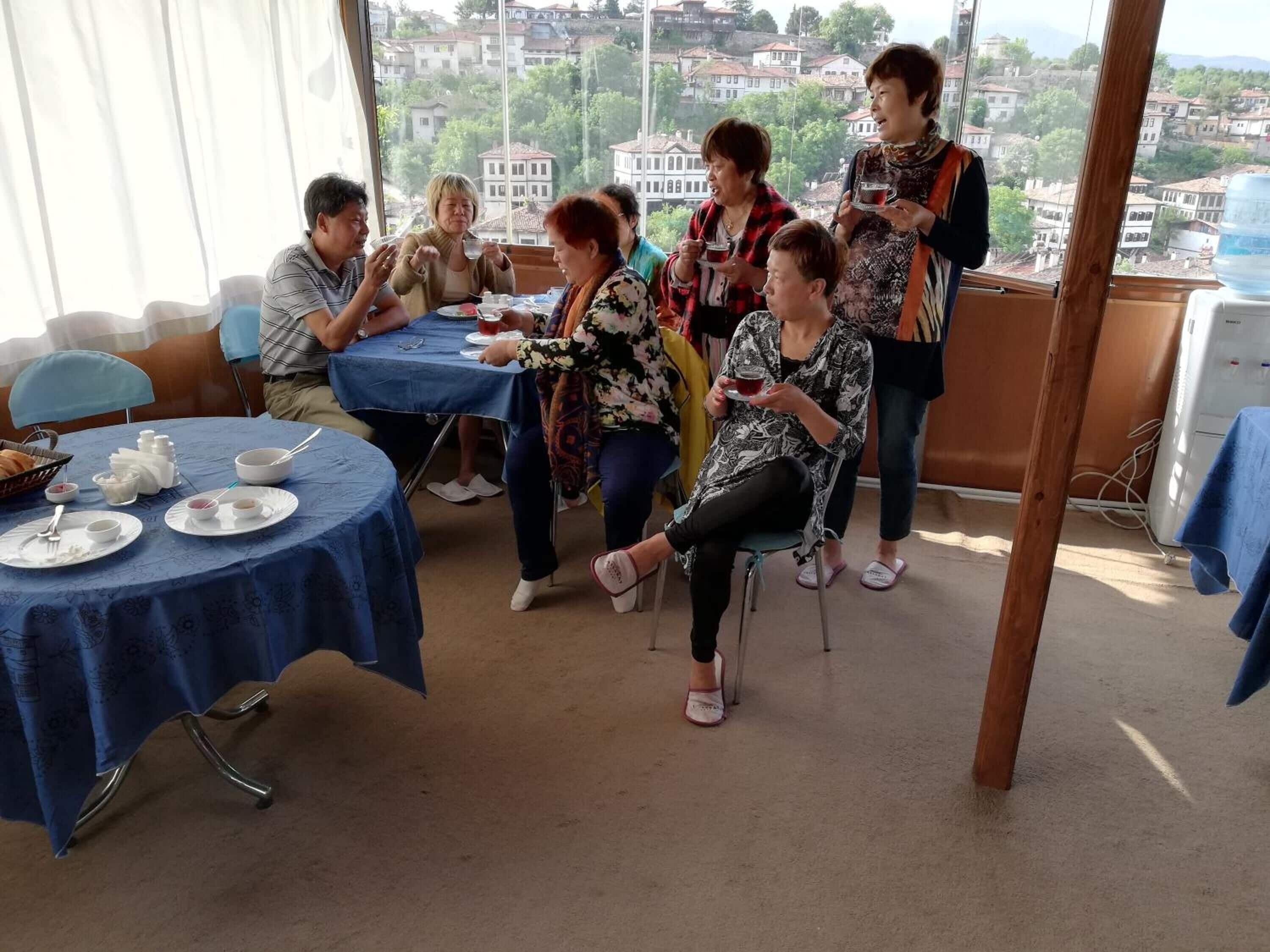 Un groupe de personnes est assis autour d’une table recouverte d’une nappe bleue, et profite d’une pause-thé tout en conversant dans une pièce d’où l’on peut admirer un paysage urbain.