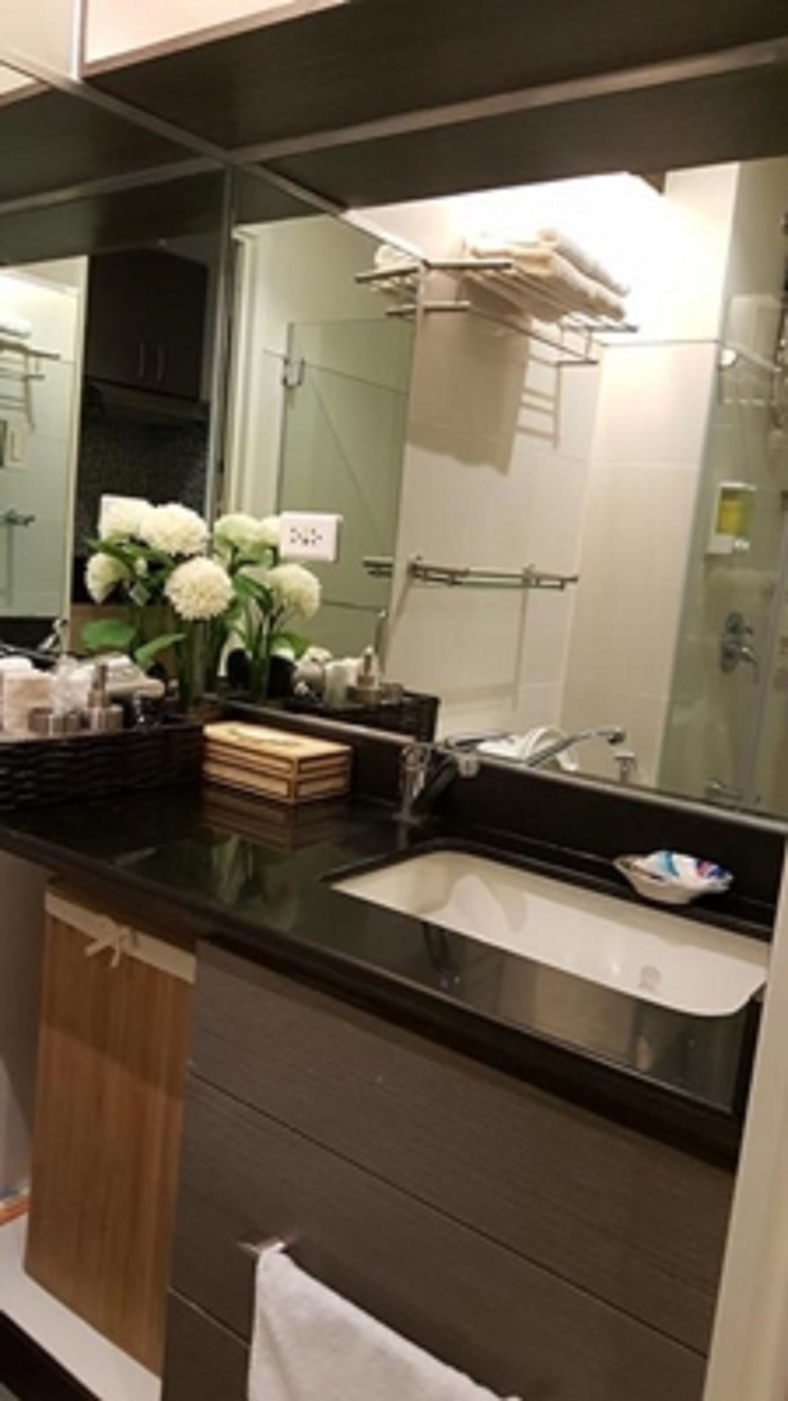 Une salle de bain moderne avec un grand miroir, un plan de toilette noir, un lavabo blanc et un vase rempli de fleurs blanches.