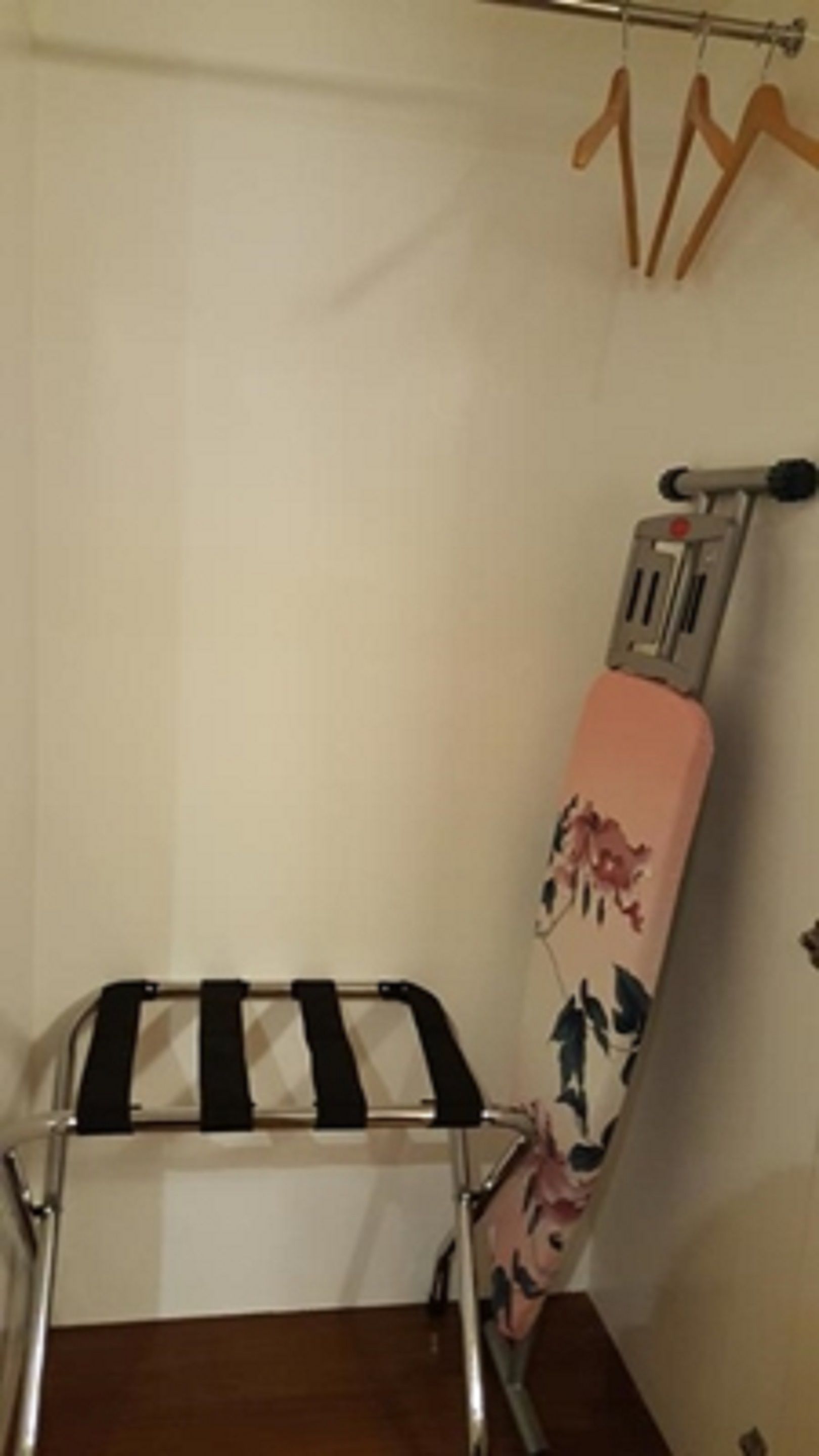 A corner of a room with a wooden floor, a wall-mounted ironing board, and a clothes rack with hangers.