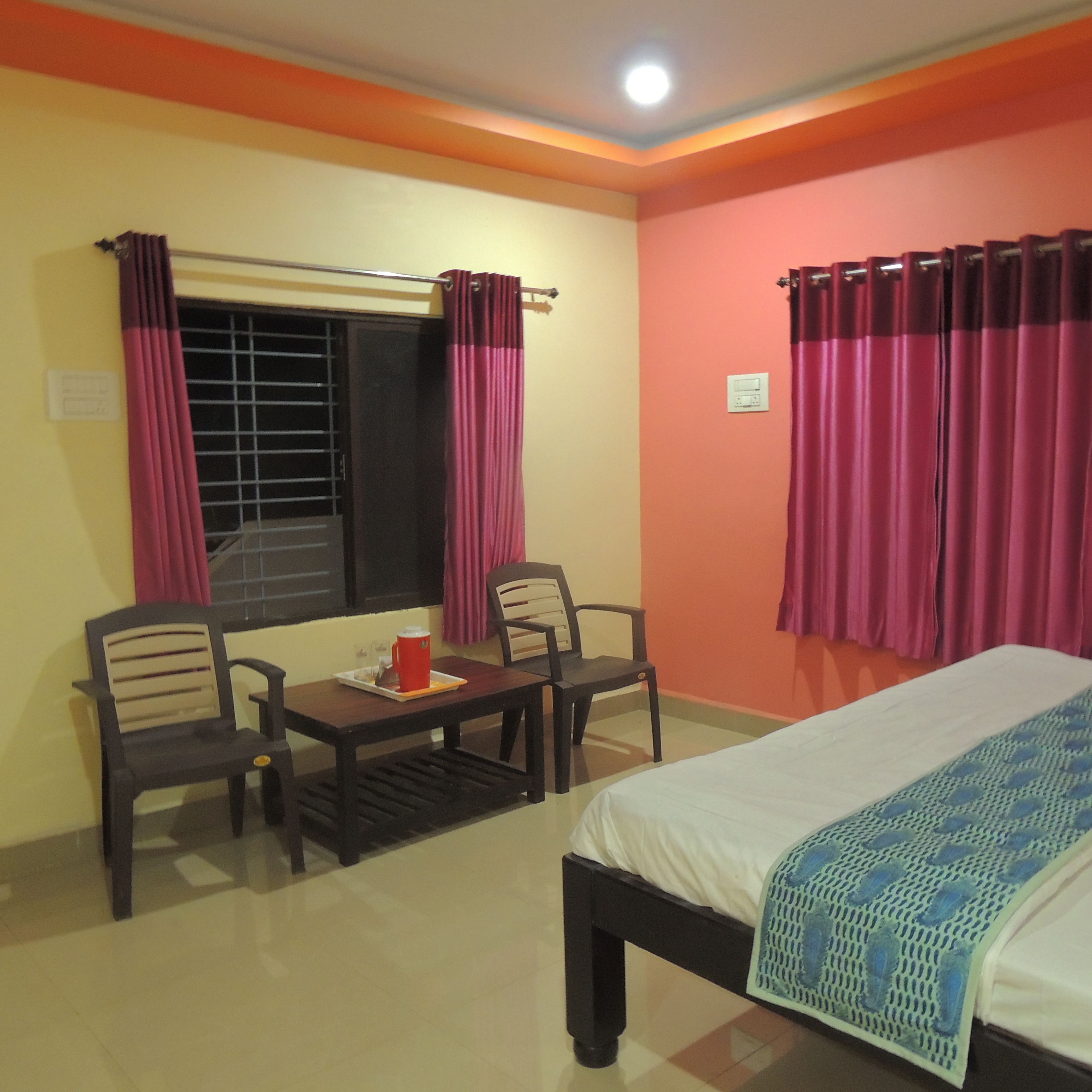 A room with a bed, two chairs, a small table with a red mug, and a window with curtains.