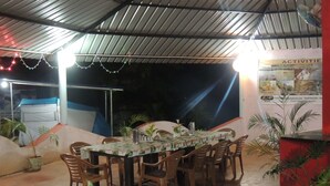 An outdoor dining area with a table set for 10, surrounded by chairs. There are potted plants and a red pillar with more plants. - Jungle Nest Homestay (Joida)