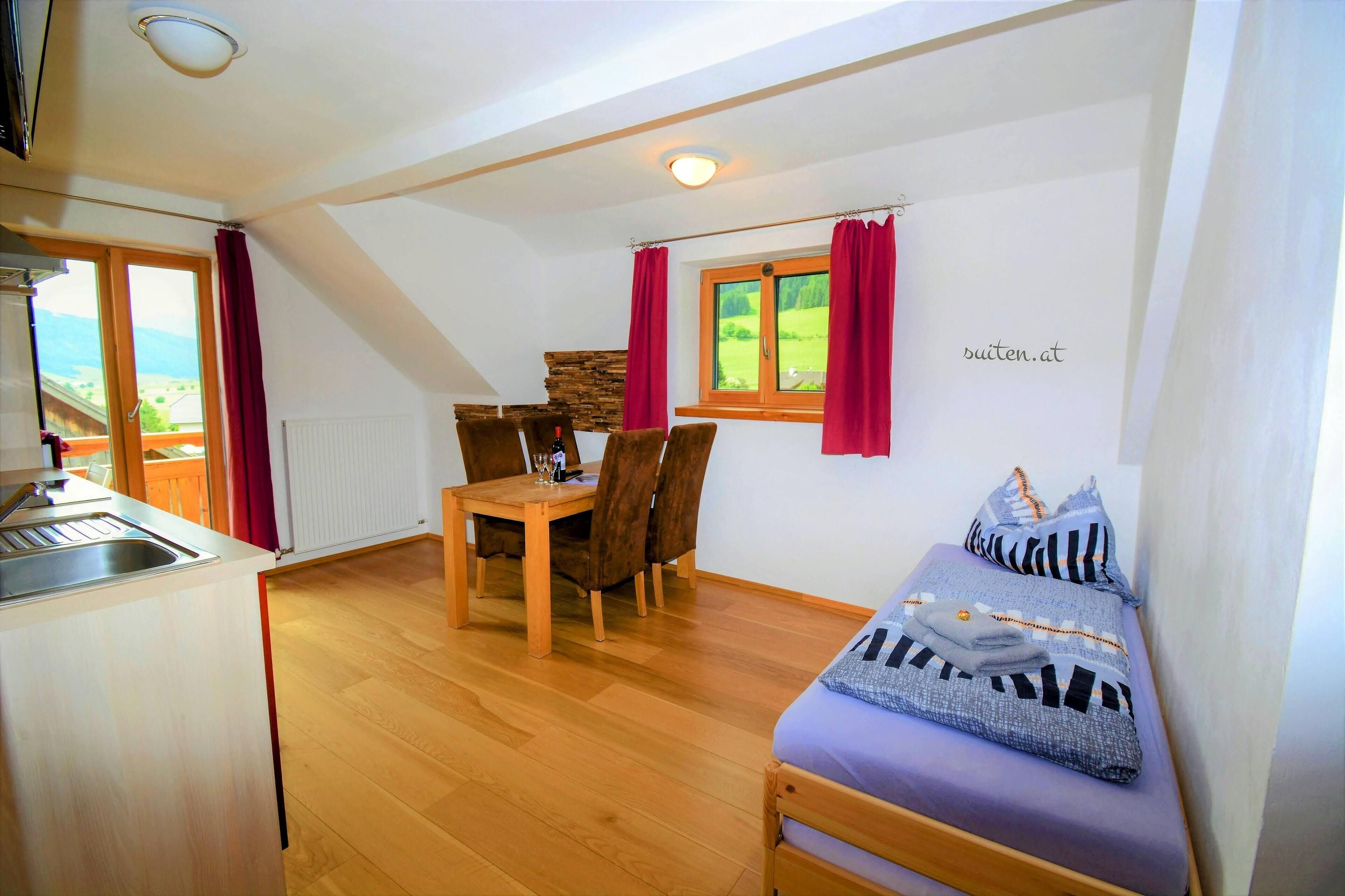 Een kleine kamer met een bed, een eettafel met stoelen, een keukenhoek en een raam met gordijnen.