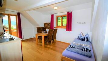 Een kleine kamer met een bed, een eettafel met stoelen, een keukenhoek en een raam met gordijnen.