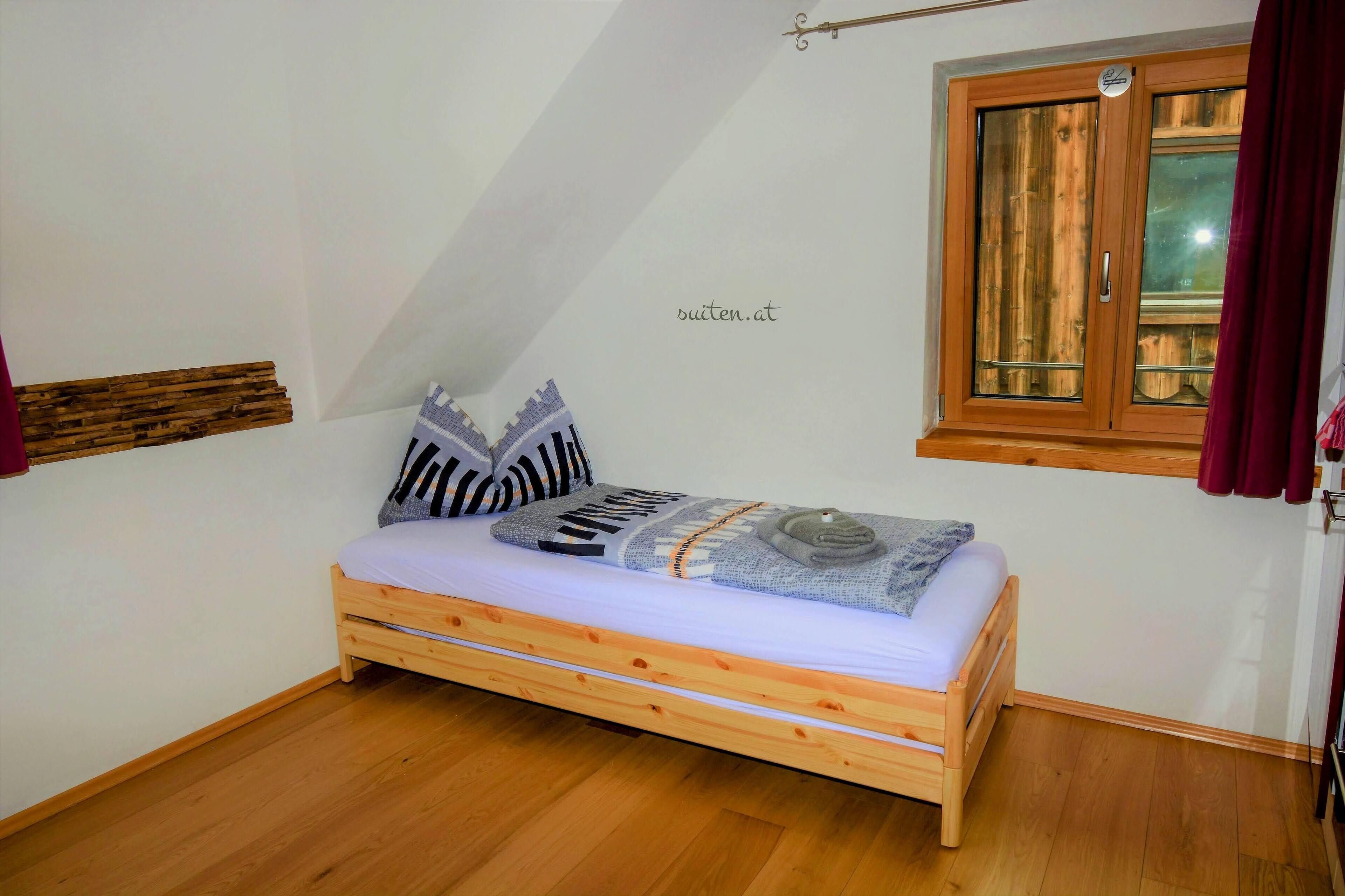 Een kleine kamer met een enkel bed, schuine wand, houten bedframe, raam met houten kader en een gordijn.