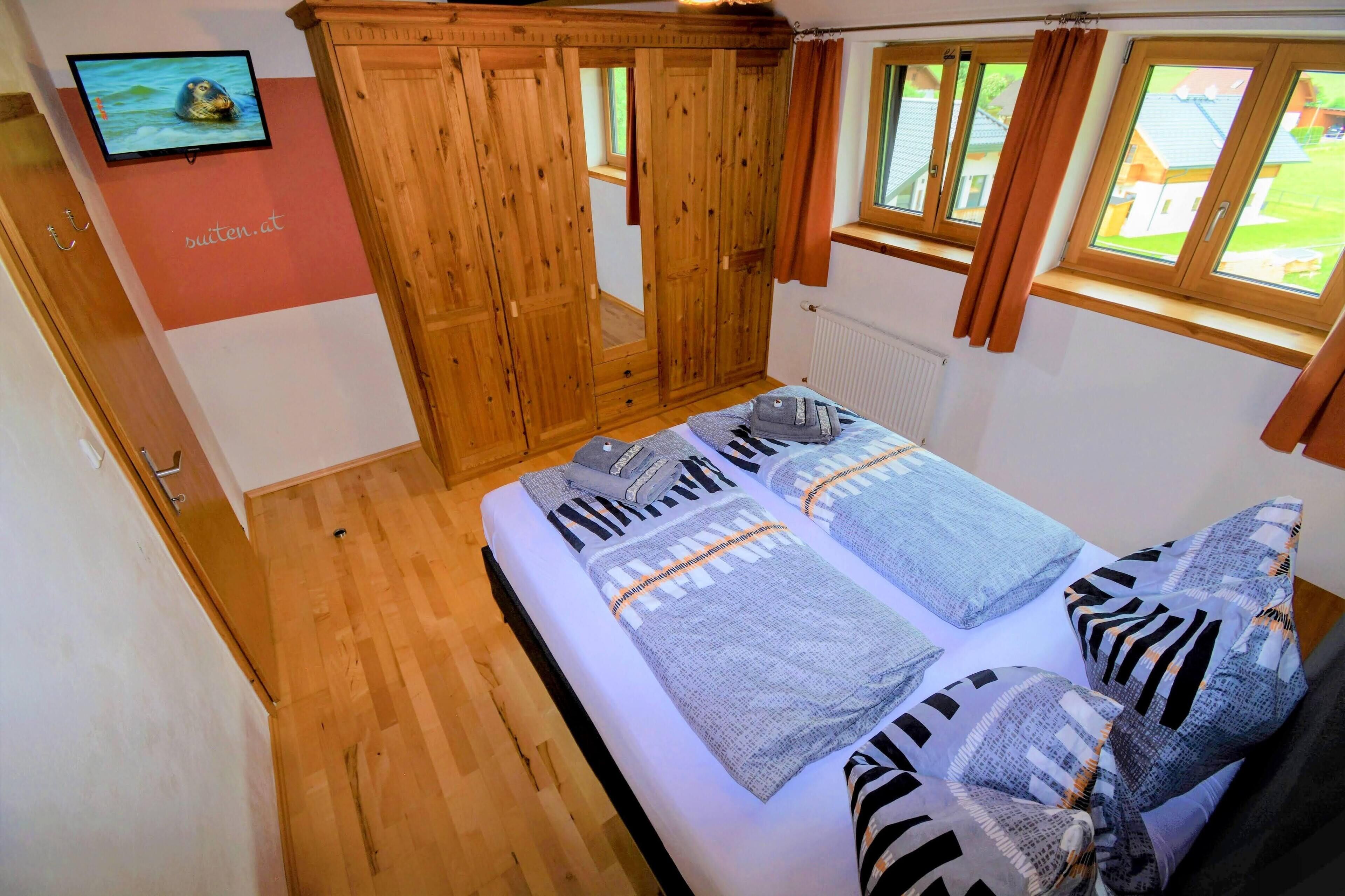 Una camera da letto con un armadio in legno, un letto con molti cuscini, una televisione a parete e una finestra con tende.