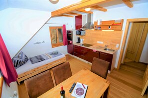 A compact kitchen with a microwave, oven, and wooden cabinets. There is a dining table with chairs and a small sofa with pillows. A red curtain hangs on the window. - David Appartments (Mauterndorf)