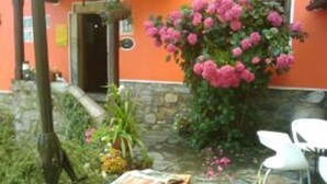 A stone patio with a wooden table and chairs, surrounded by lush greenery and pink flowers.