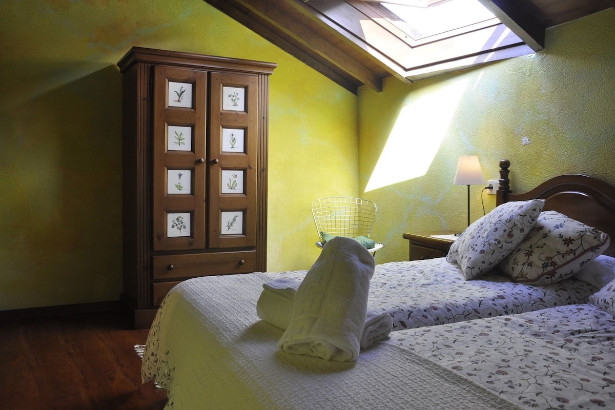 A bedroom with a skylight, a wooden cabinet with framed botanical prints, a bed with floral bedding, and a nightstand with a lamp.