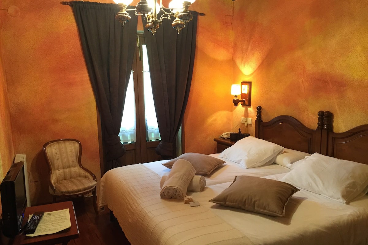 A hotel room with a large bed, a chair, a desk with a remote control and papers, and a wall-mounted lamp.