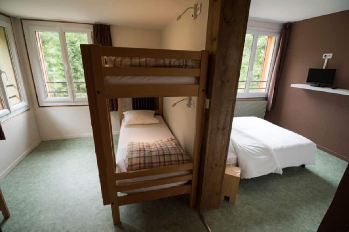 A room with bunk beds, a small desk, a television on a shelf, and windows with curtains.