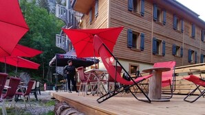 A wooden deck with red umbrellas, outdoor seating, and a large log. - 1861 Châtel Hostel (Chatel)