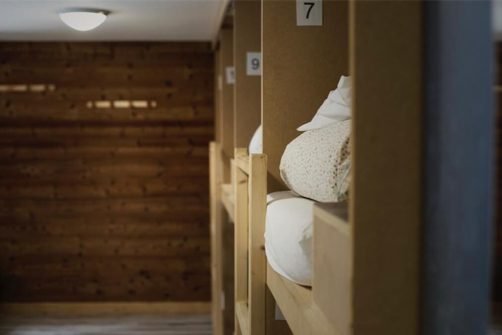 Filas de literas de madera en una habitación con techo inclinado y paredes de madera. Cada cama tiene un número en la pared superior.