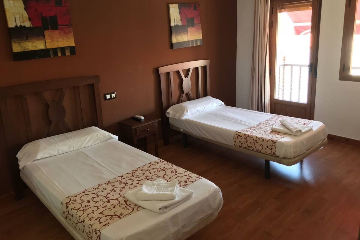 A hotel room with two single beds, wooden headboards, a small bedside table with a lamp, and a window with curtains.