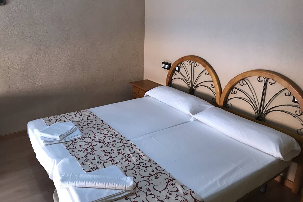 A neatly made bed with a patterned bedspread and white pillows, a wooden nightstand, and a wall-mounted light switch.