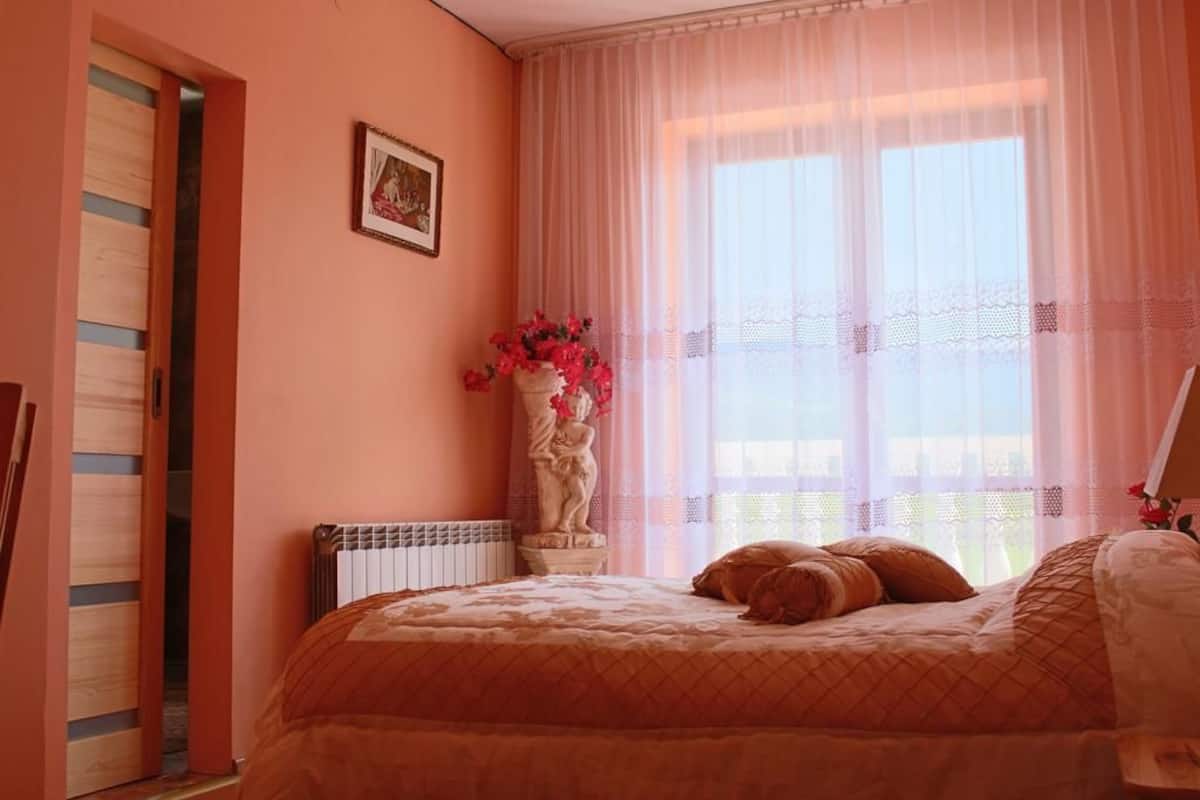A bedroom with a bed, a radiator, a statue, and a window with sheer curtains.