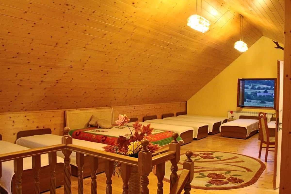 A cozy room with a sloped ceiling, wooden walls, and a large window. There are three beds arranged in a row, a wooden table with a vase of flowers, and a chair.