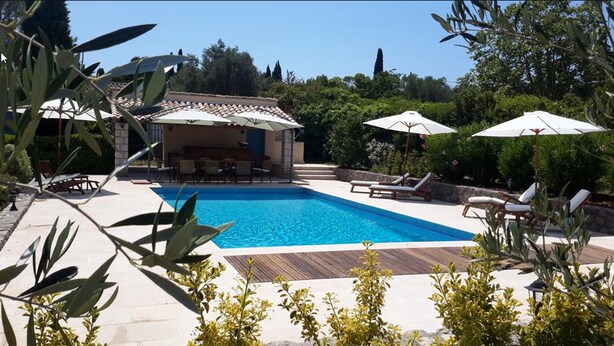 Una piscina con sillas de descanso y sombrillas, rodeada de vegetación y un edificio con mesas al aire libre.