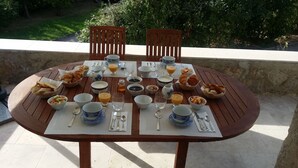 Mesa de comedor al aire libre preparada con productos de desayuno, incluyendo cruasanes, fruta y bebidas, rodeada de sillas.