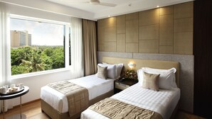 Una habitación de hotel con dos camas, una mesita con una bandeja de desayuno, una mesita de noche con lámpara y un ventanal con vistas a la ciudad.