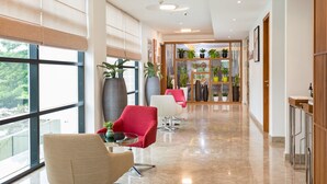 Un moderno hall de hotel con un largo pasillo, un área de descanso con sillas rojas y beige, grandes macetas y vistas al exterior a través de amplias ventanas.