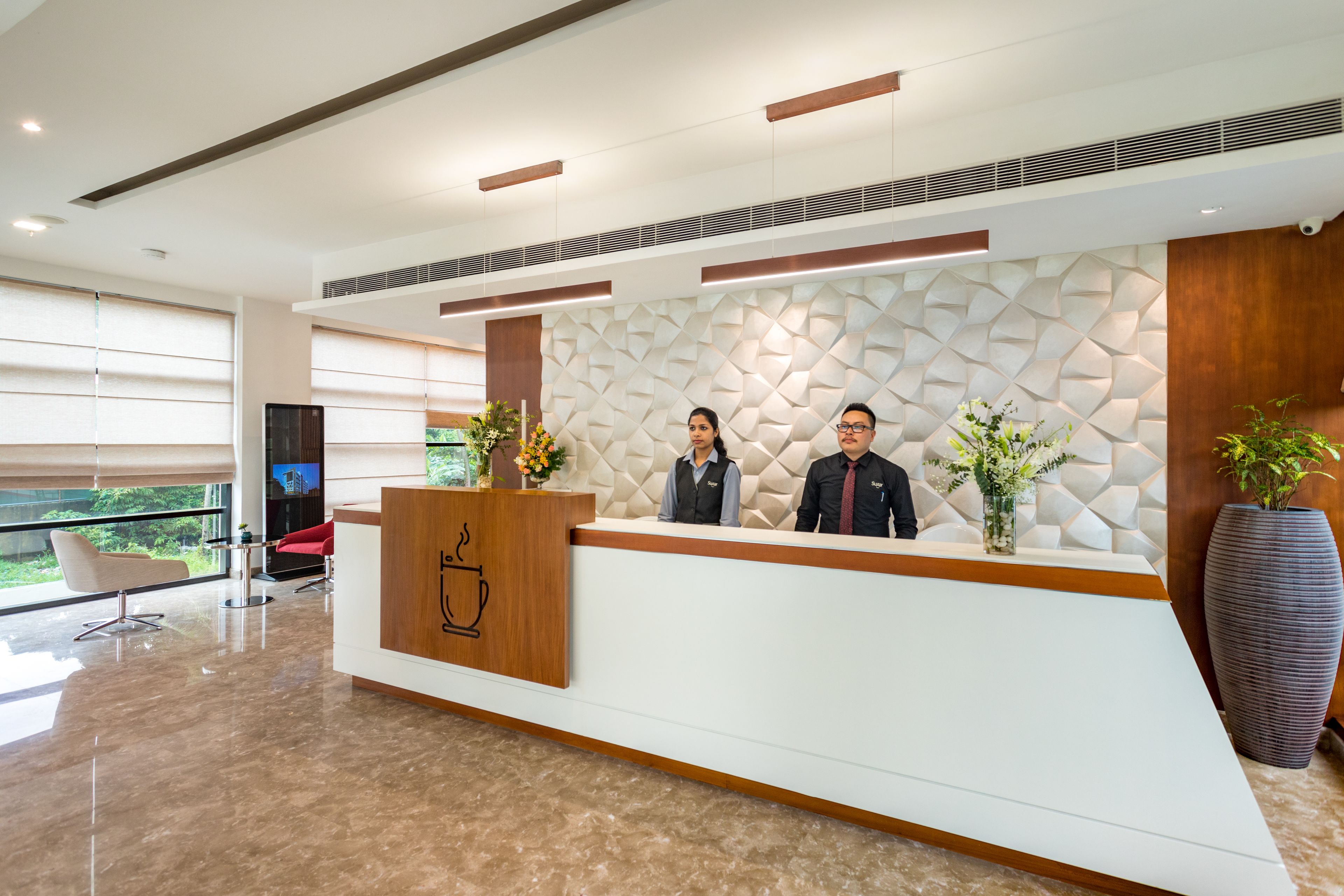 A modern reception desk with two staff members, a large potted plant, and a view of the outdoors through large windows.