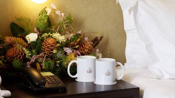 Ein Hotelzimmer mit einem Nachttisch, der eine Blumenkomposition, zwei weiße Tassen mit einem Logo und ein schwarzes Telefon zeigt.