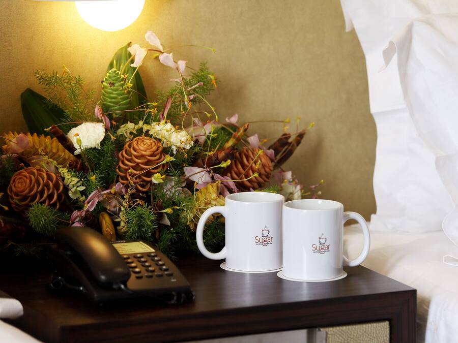 Ein Hotelzimmer mit einem Nachttisch, der eine Blumenkomposition, zwei weiße Tassen mit einem Logo und ein schwarzes Telefon zeigt.