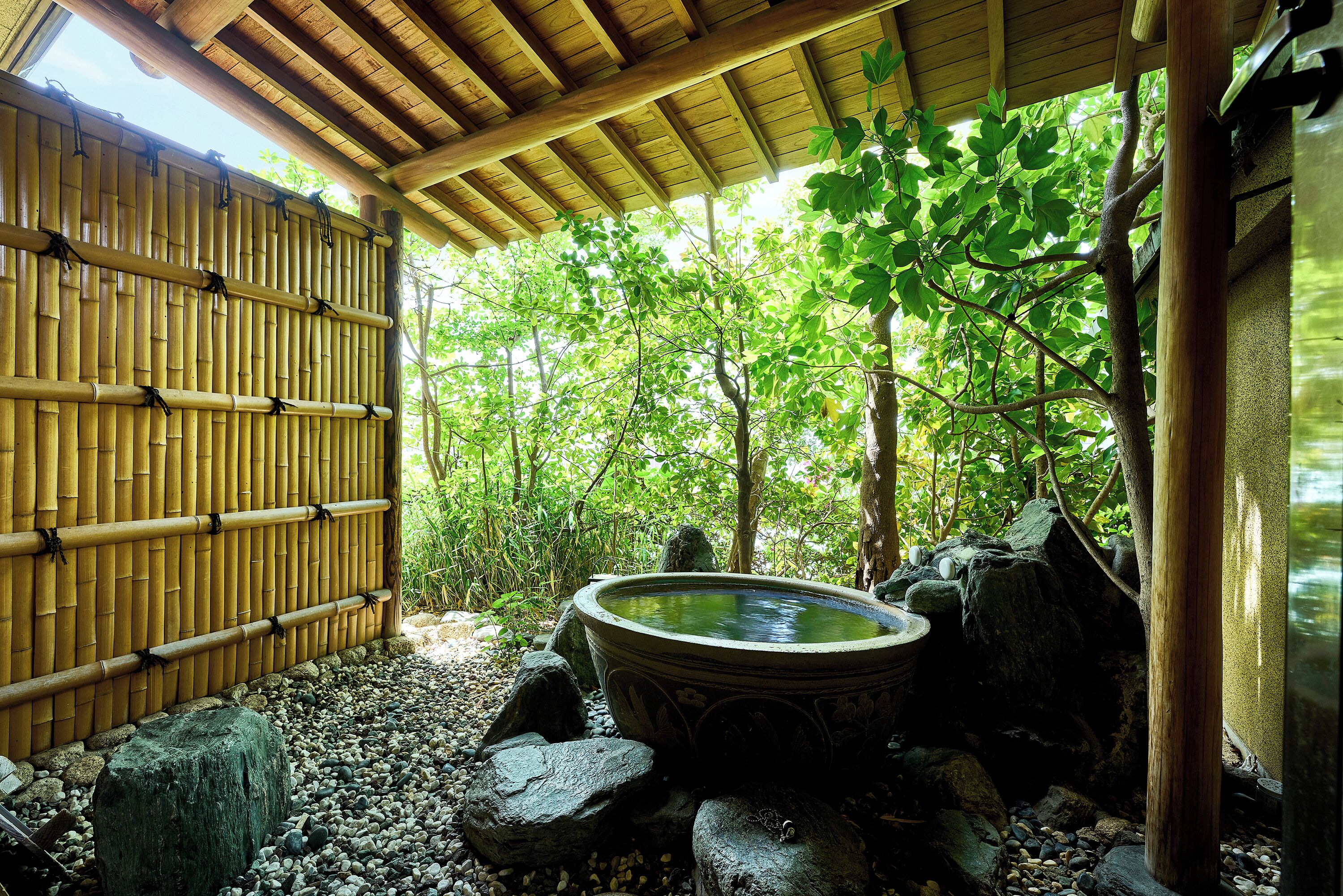 Un jardin japonais traditionnel avec un bain à remous en pierre, un mur de bambou et une pergola en bois.