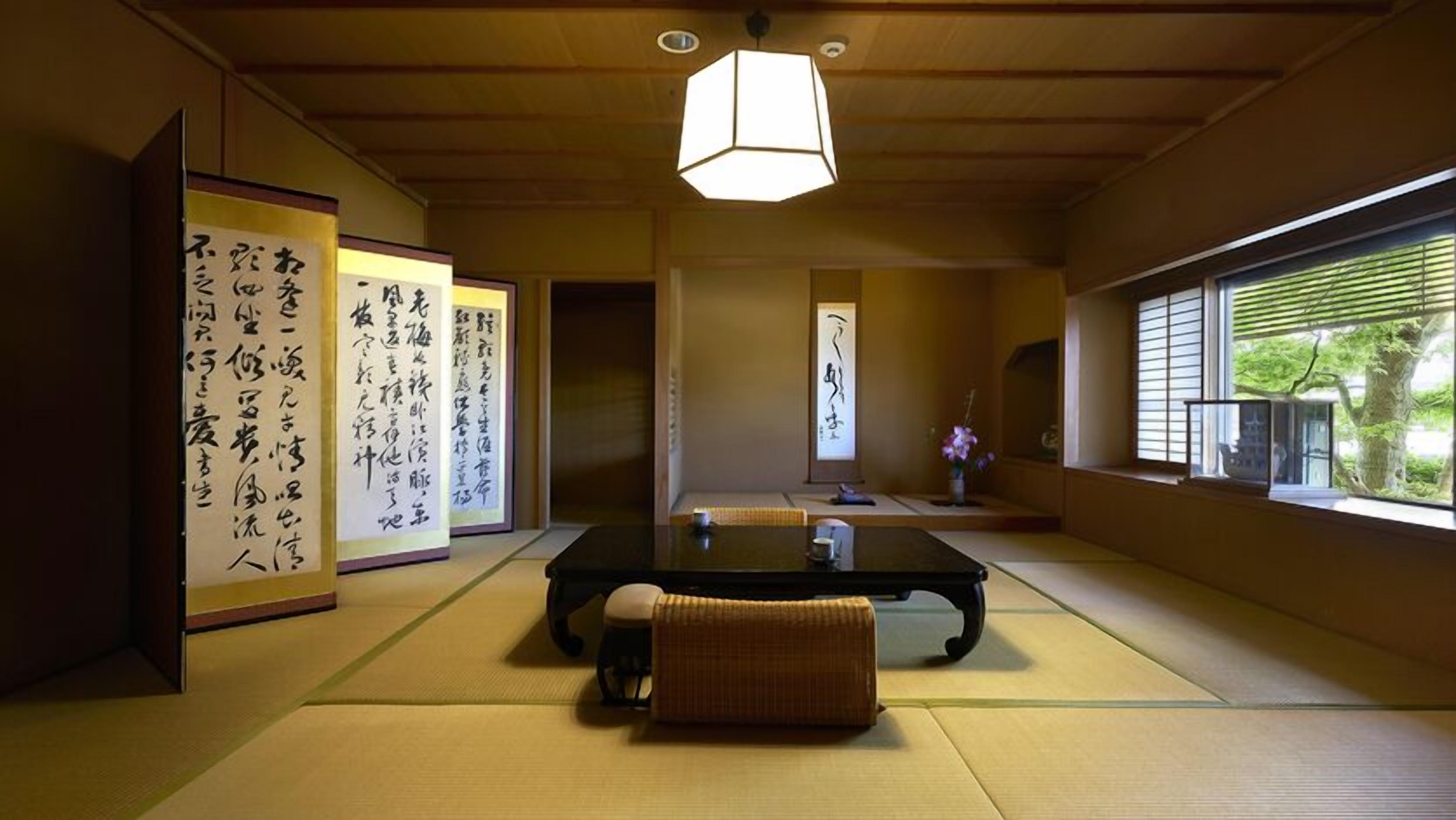 Una habitación japonesa tradicional con suelo de tatami, una mesa baja, un banco acolchado, rollos de caligrafía y vistas a través de una ventana.
