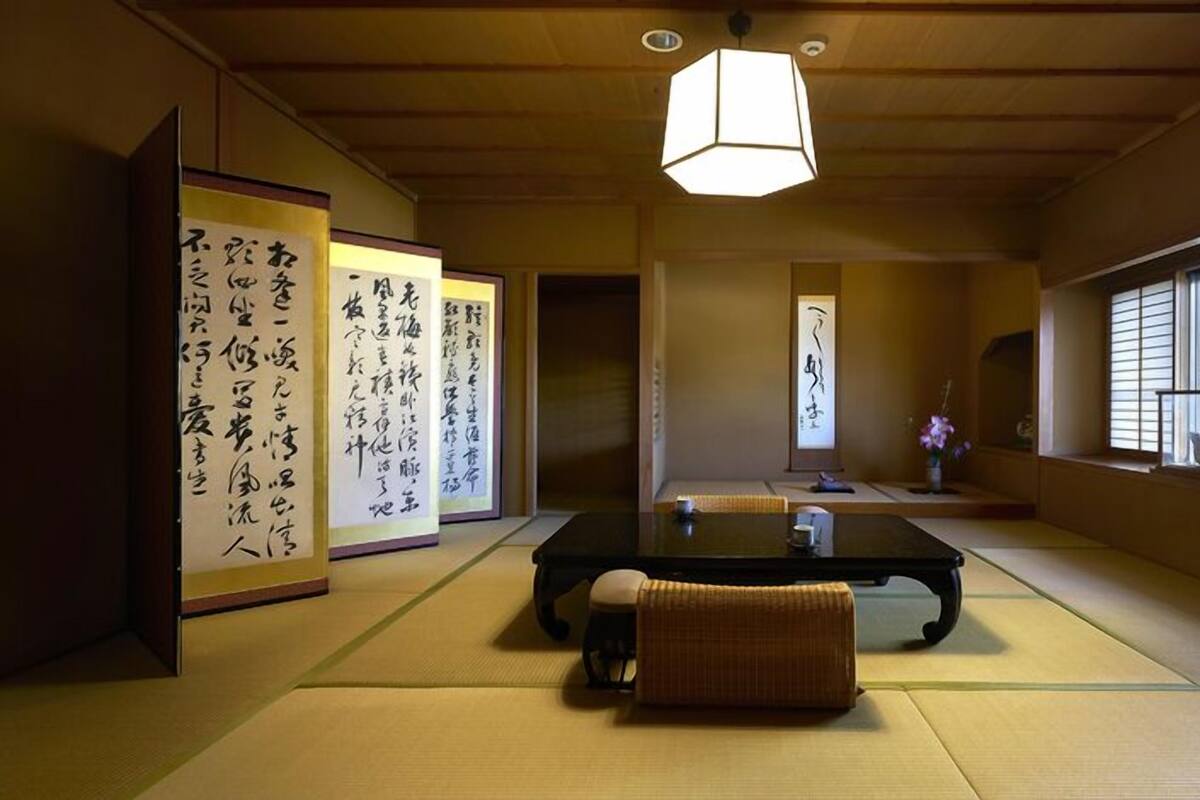 Um quarto japonês tradicional com tatames, uma mesa baixa, um banco almofadado, rolos de caligrafia e vista pela janela.