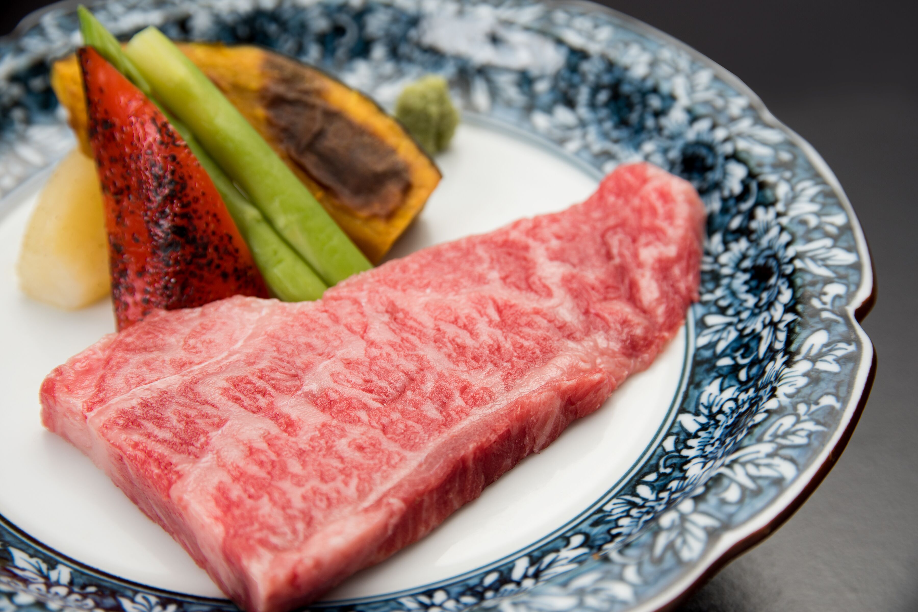 A plate with a piece of raw meat, vegetables, and a patterned bowl.
