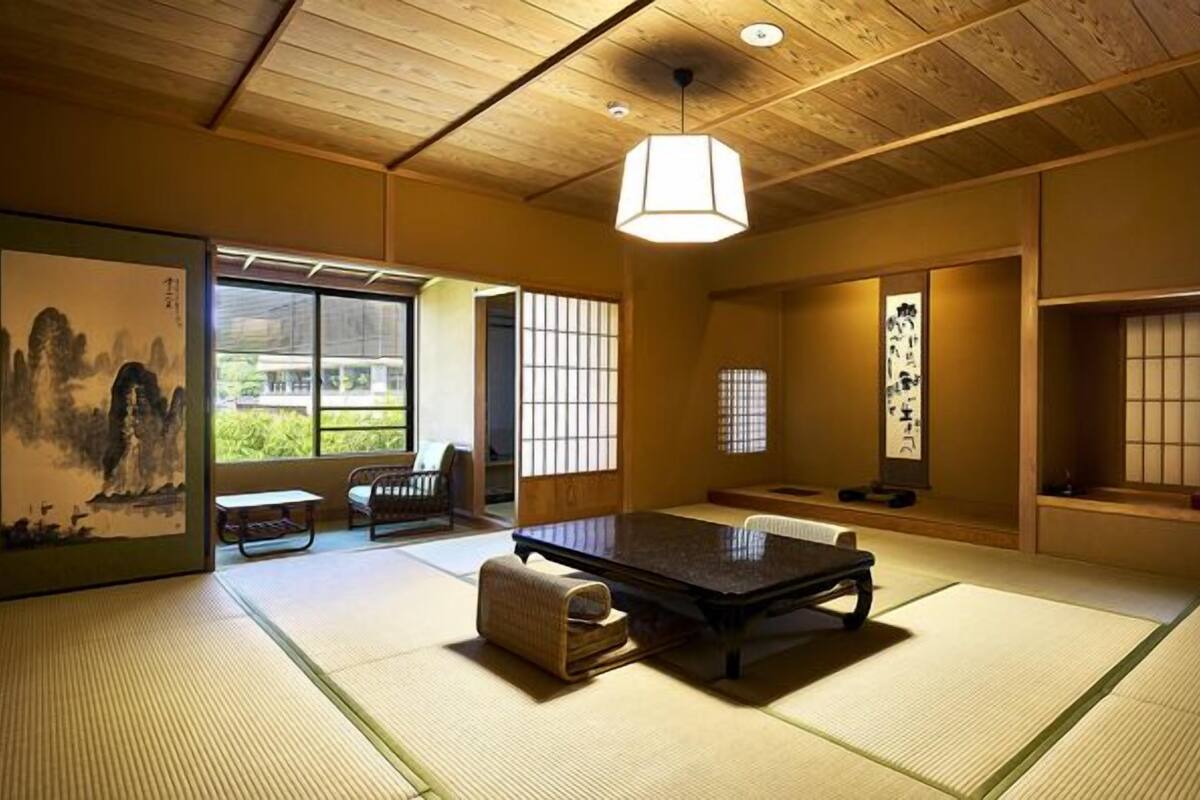 Um quarto japonês tradicional com piso de tatame, uma mesa baixa, luminária pendente, portas deslizantes e vista para o verde lá fora.