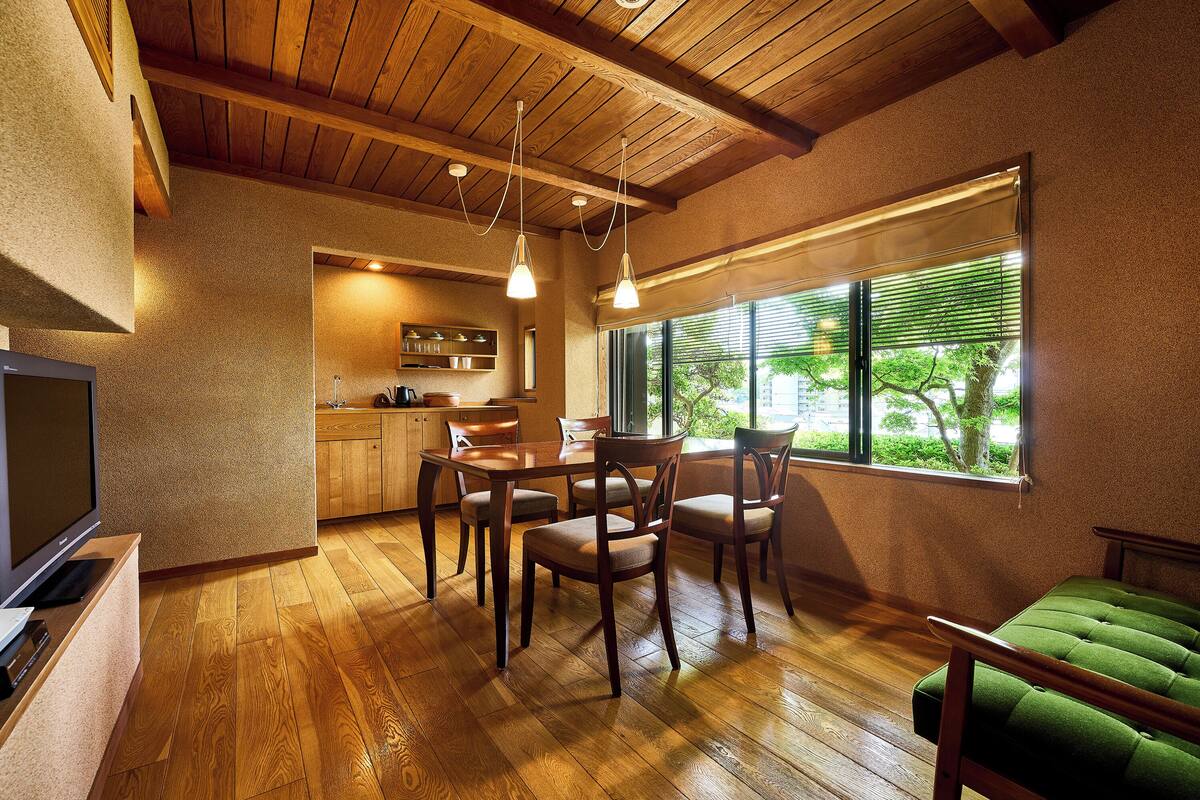 Uma sala de jantar com piso de madeira, mesa e cadeiras, e vista para o exterior através de uma janela.