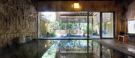 A traditional Japanese onsen with a stone wall, a wooden ceiling, and a view of a garden through glass doors.